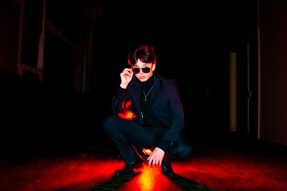 Man in dark suit adjusting sunglasses while crouching over red light on dark floor.