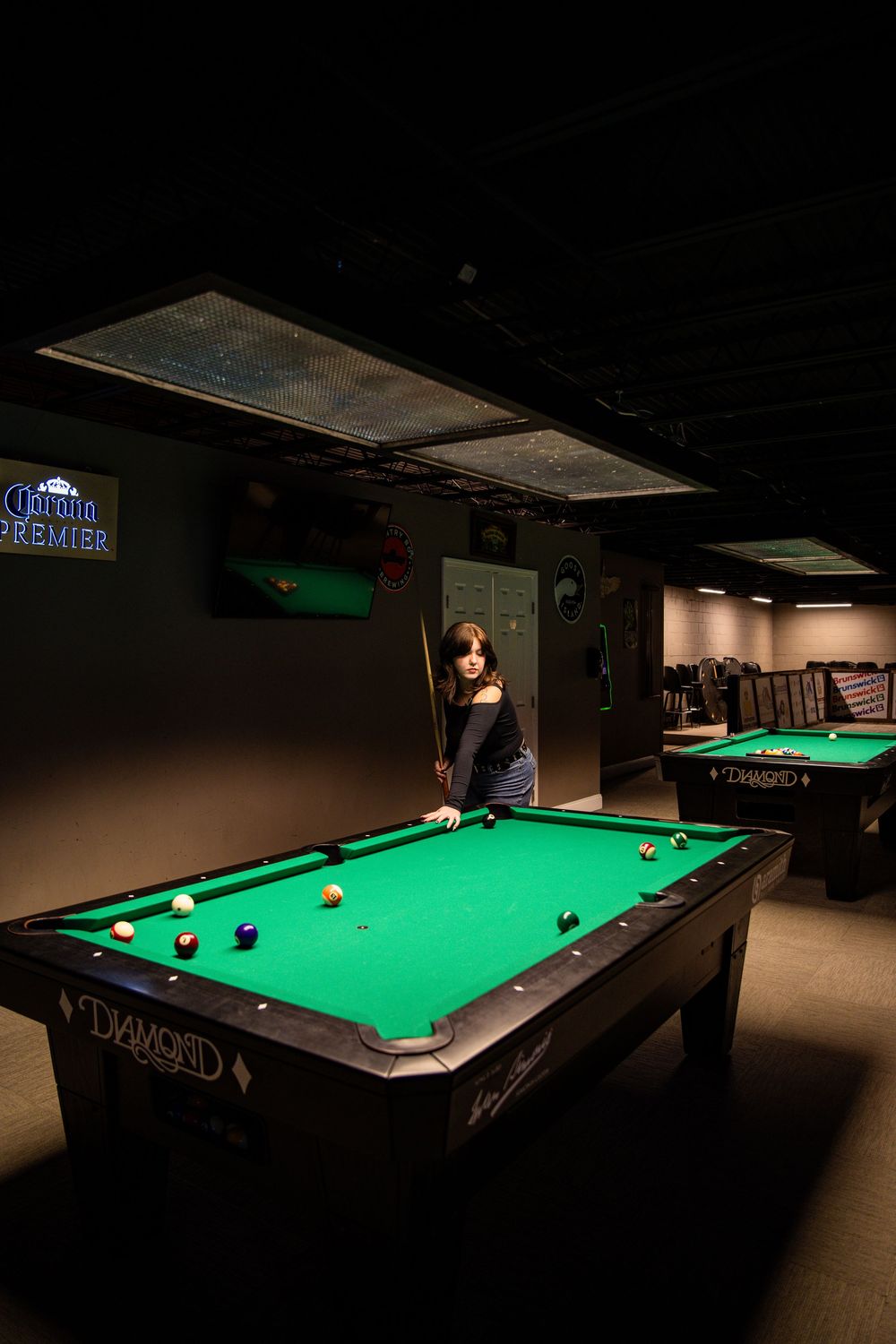 Woman playing pool on a Diamond billiard table in a dimly lit bar with Corona Premier neon sign.