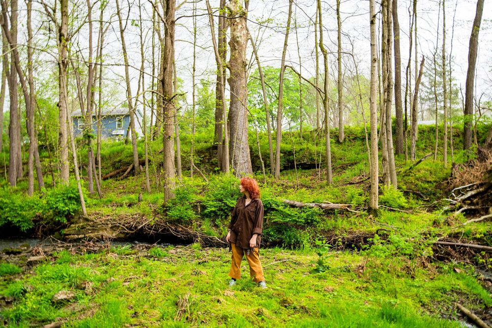 Red-haired woman standing in a lush green forest with tall trees and mossy ground.