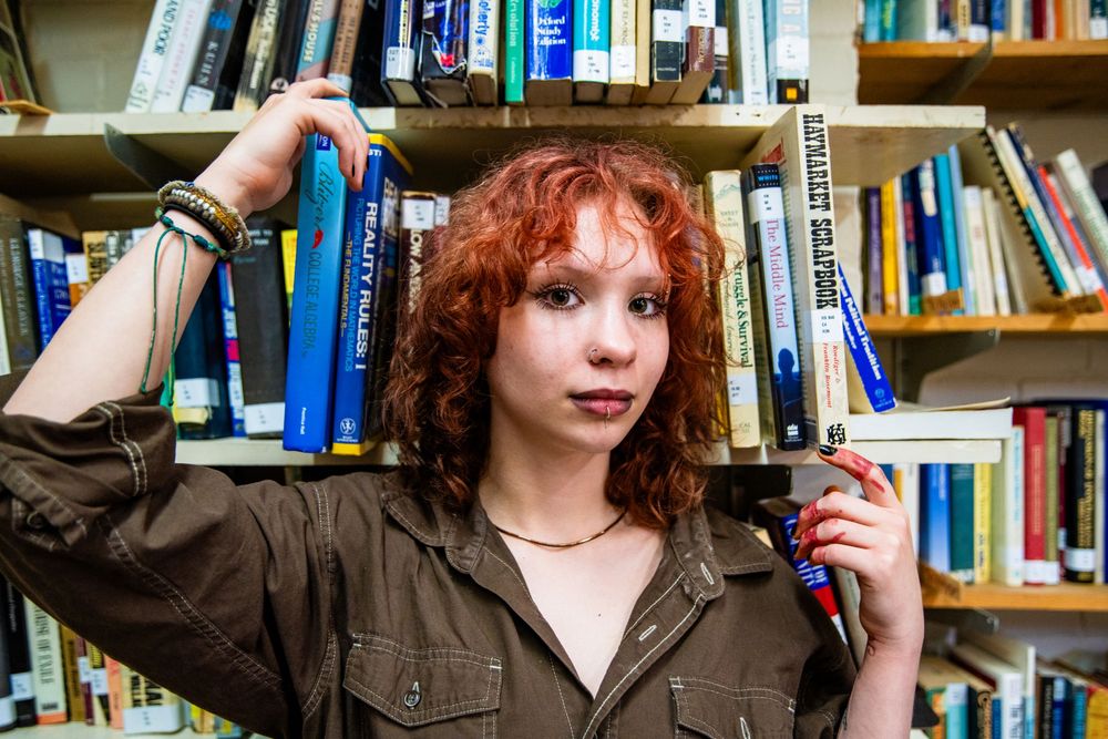 Curly red-haired young woman posing between bookshelves in a library holding books.