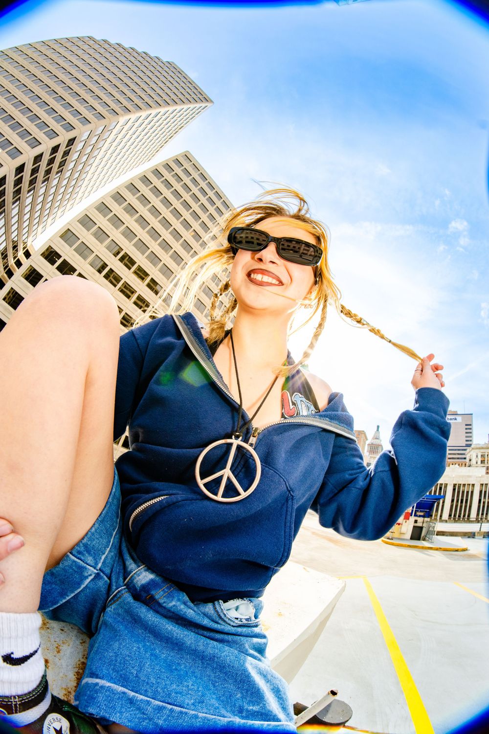 Smiling blonde woman in blue hoodie and peace sign necklace poses on rooftop with city skyscrapers behind her.