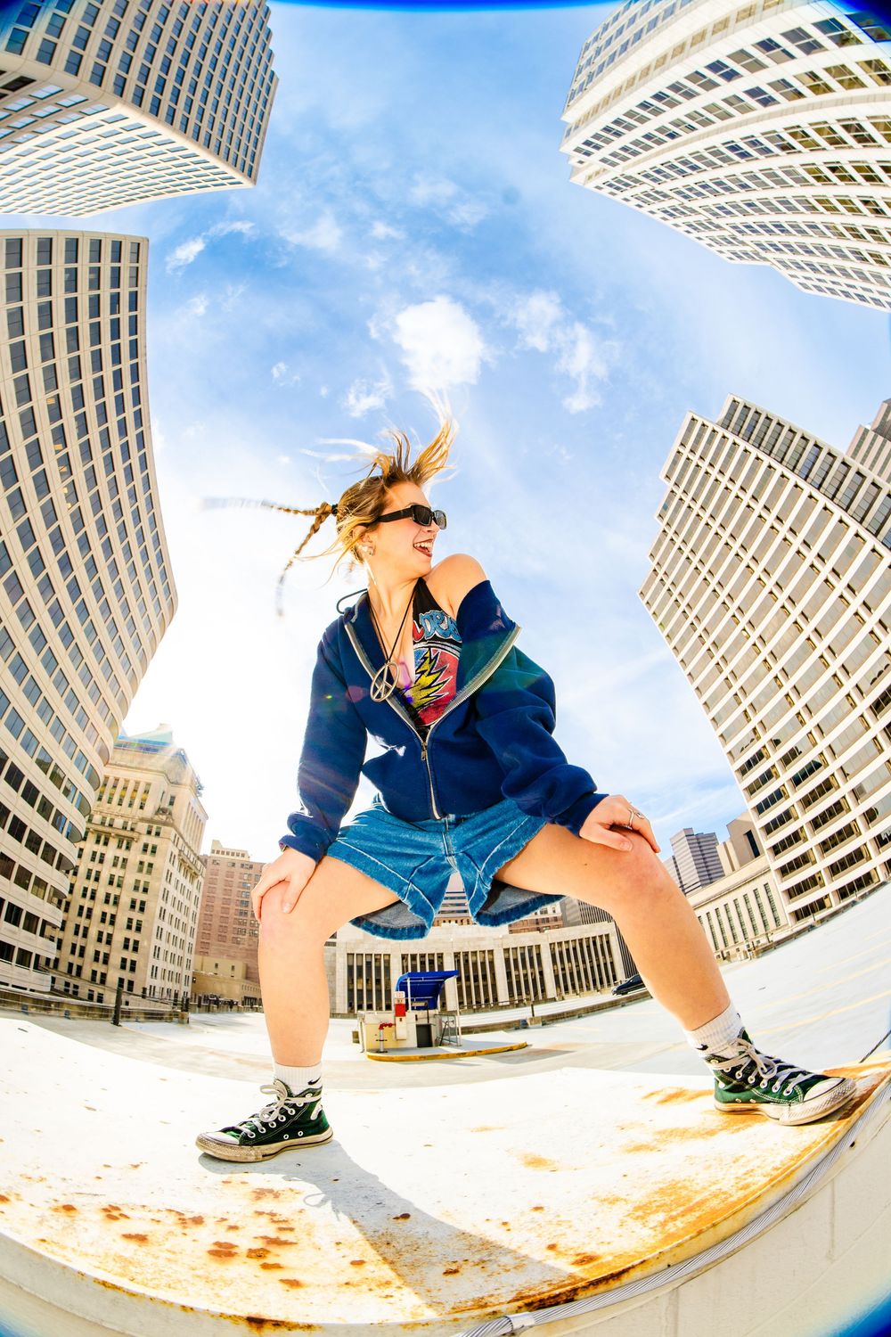 Energetic blonde woman in blue hoodie and denim shorts poses dynamically on rooftop surrounded by tall city buildings.
