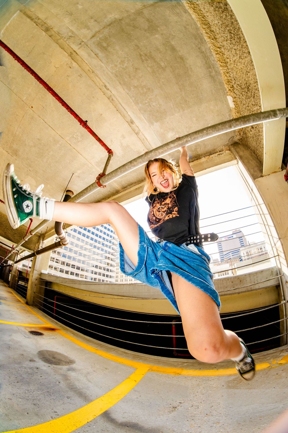 Blonde woman in black graphic tee and denim overalls kicks leg high in urban parking garage with fisheye lens.