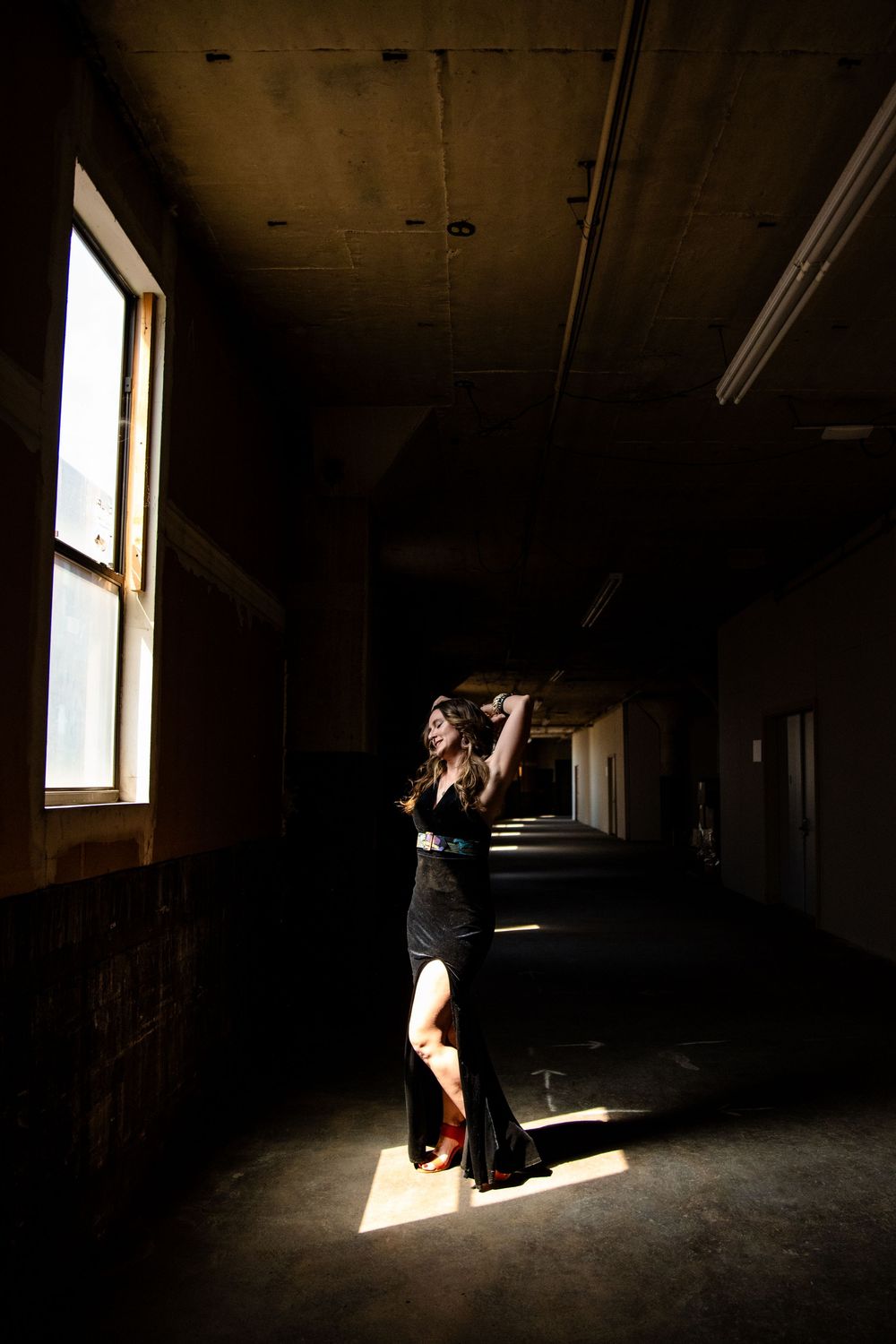 Woman in black dress posing dramatically in a dark, abandoned hallway with a single beam of natural light.