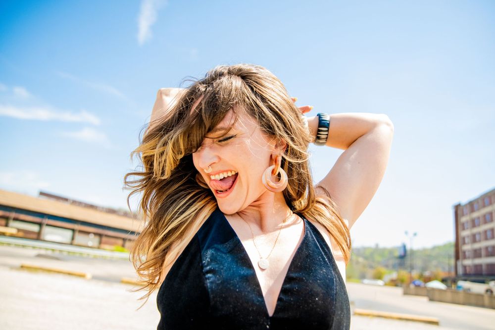 Woman with flowing blonde hair laughing outdoors on a sunny day, wearing a black top and hoop earrings.