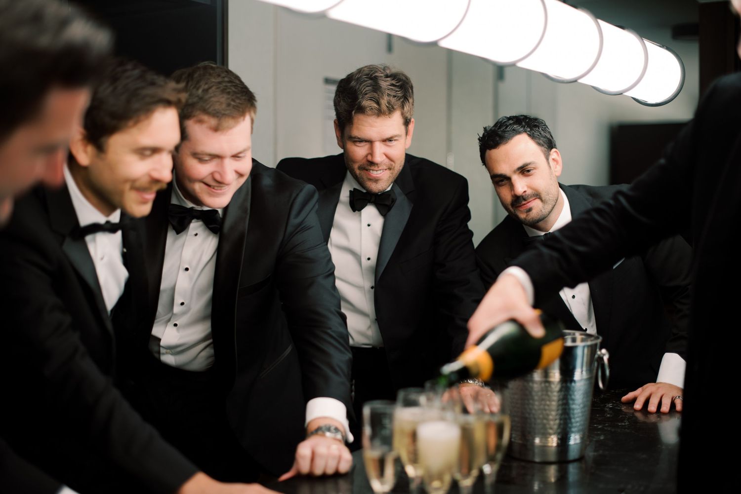 Four men in tuxedos celebrate while pouring champagne at a bar, smiling and enjoying a festive occasion.