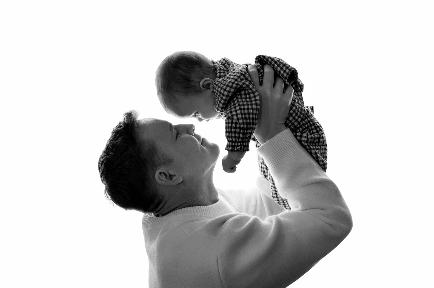 Black and white photo of a father joyfully lifting his smiling baby up in the air against a bright white background.