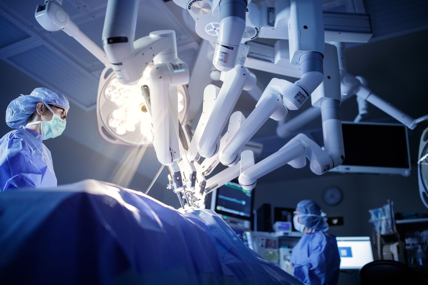 Robotic surgical system with da Vinci robot arms operating on a patient while surgeons in blue scrubs monitor the procedure.