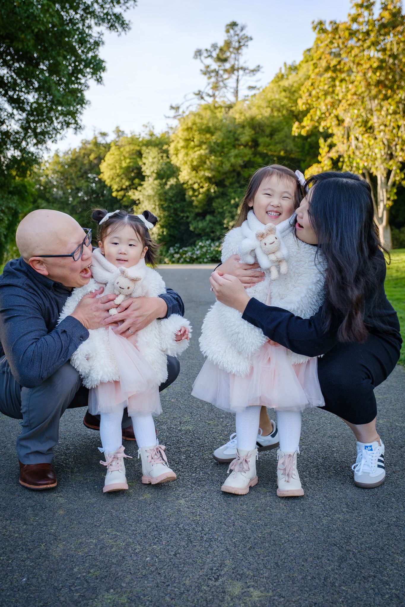 Family of four with twin girls in pink tutus and white fur coats, posing outdoors with parents in a park.