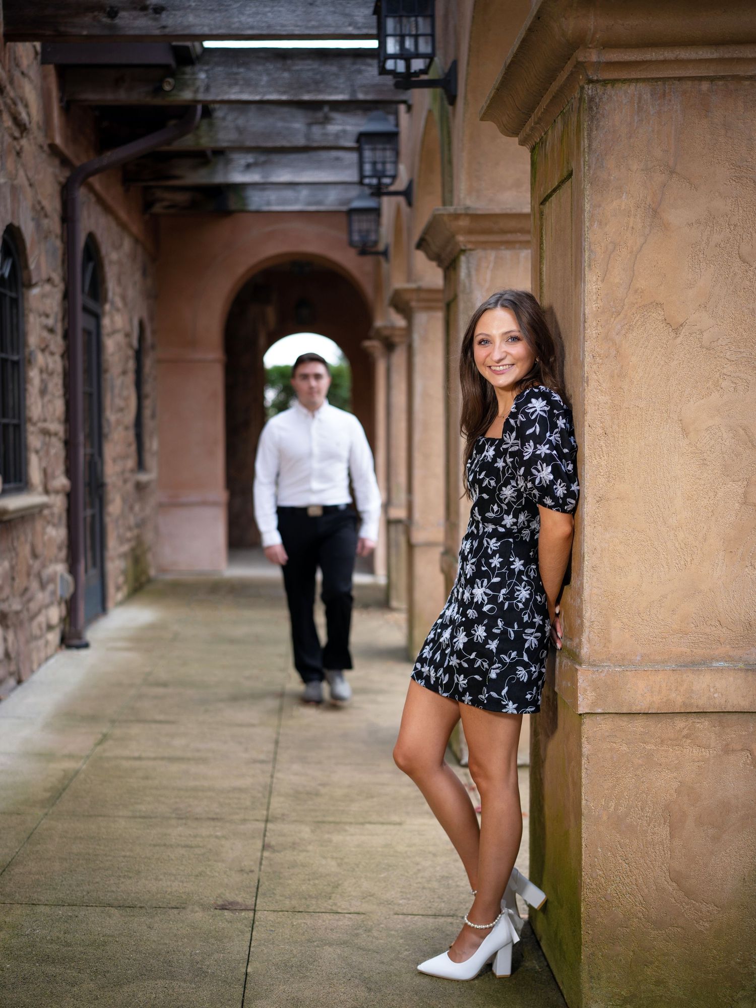 Young woman in floral dress leaning against stone column smiling, man in white shirt walking through arched corridor behind her.