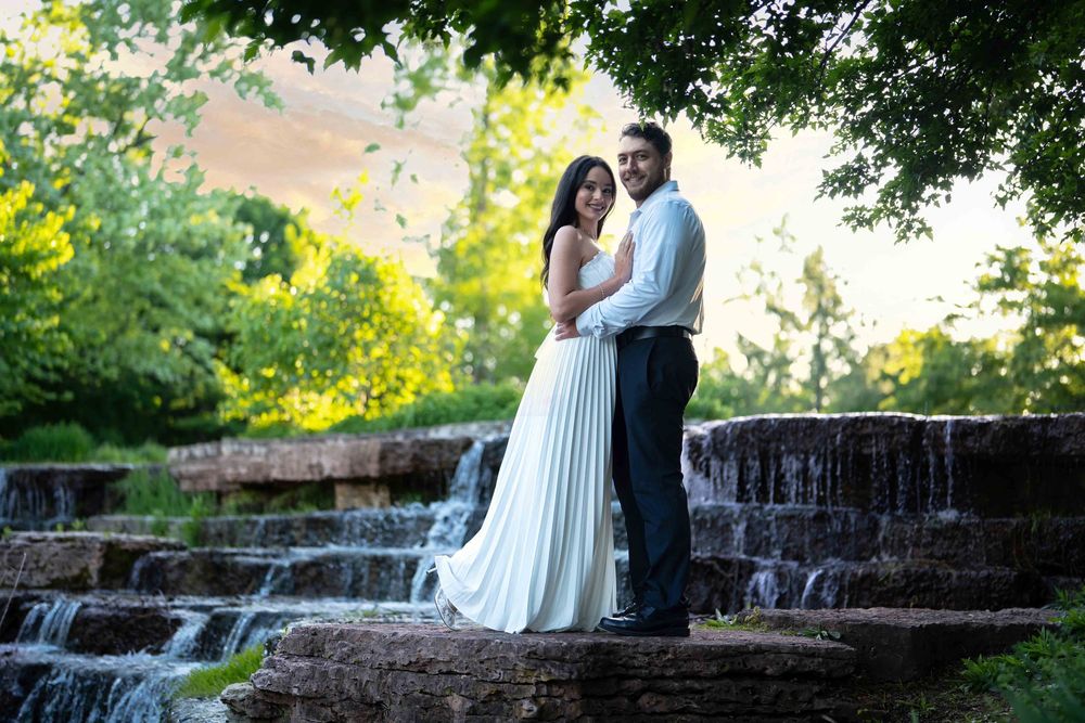 Bride and groom pose romantically by a stone waterfall surrounded by lush green trees in an outdoor wedding photo.