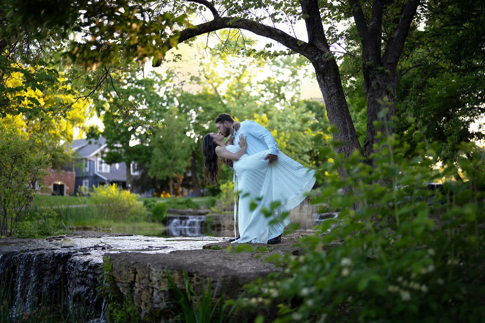 Bride and groom share a romantic dip kiss surrounded by lush green trees near a garden waterfall.