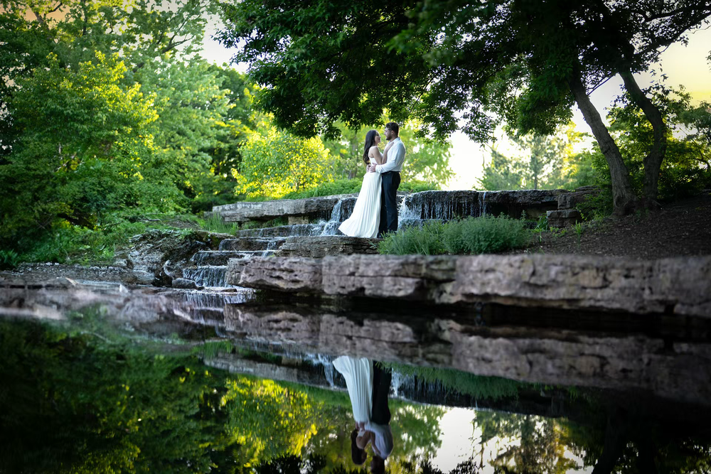 Bride and groom embracing near a serene waterfall and pond in a lush green garden setting.