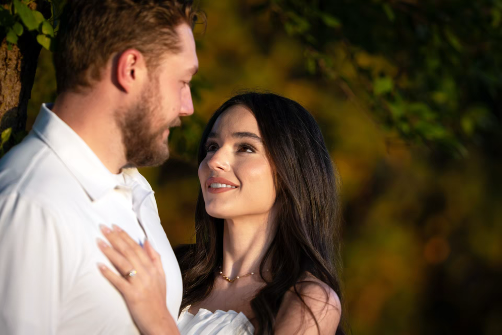 A couple shares a romantic moment outdoors, woman smiling up at bearded man against golden autumn foliage backdrop.