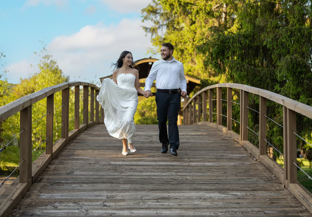 Couple walking hand-in-hand on a wooden bridge, woman in white dress, man in white shirt, surrounded by lush greenery.