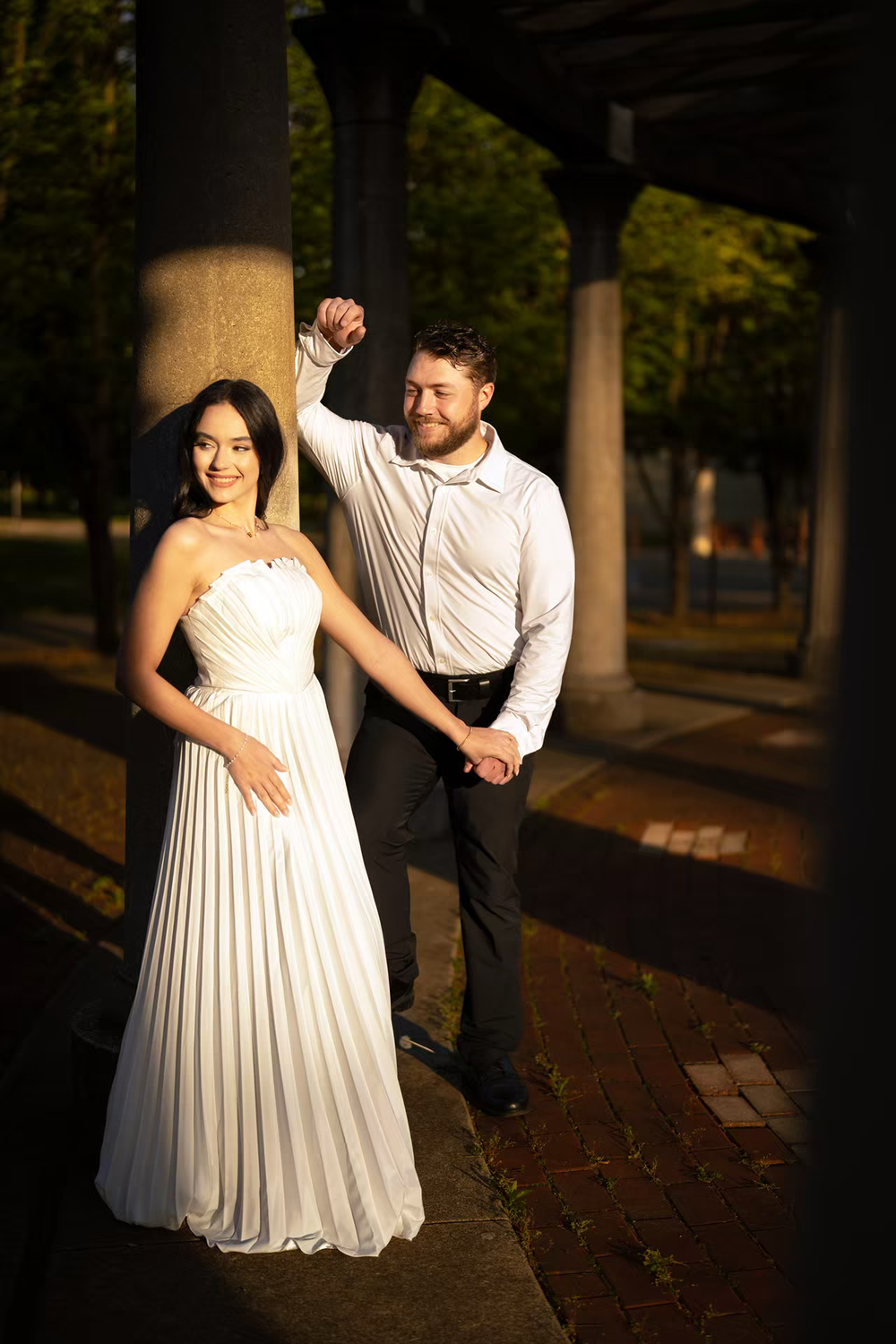 A smiling couple in wedding attire poses near stone columns in a sunlit park, groom in white shirt and bride in elegant white gown.
