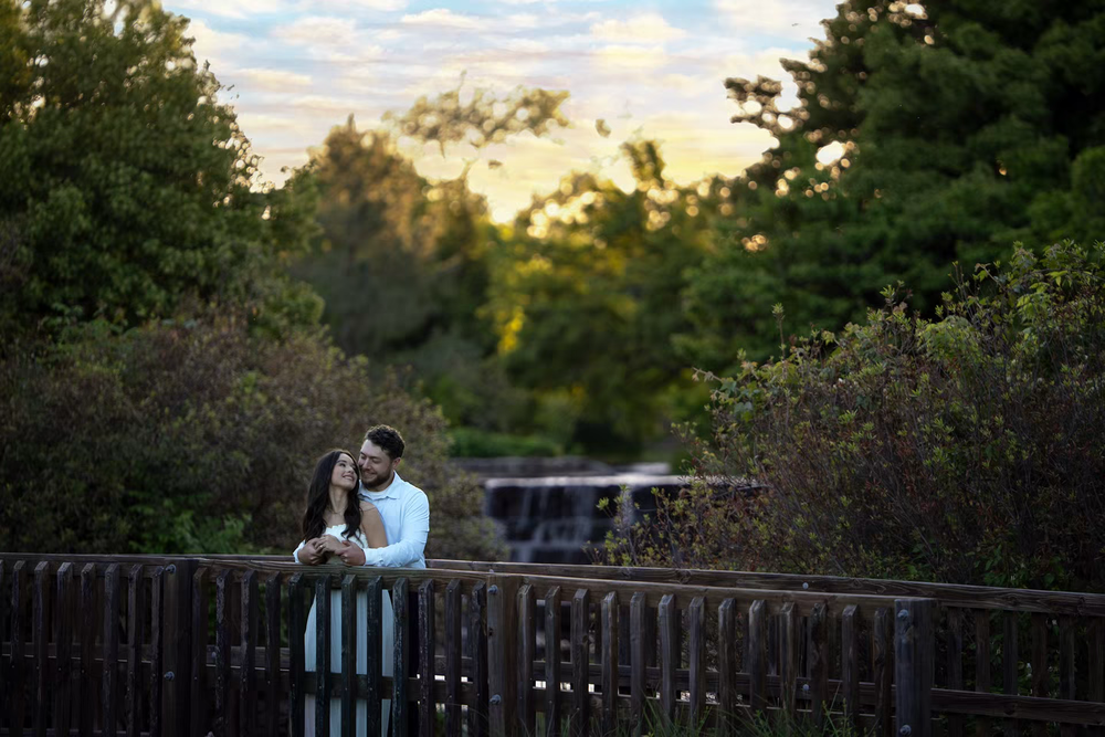 Couple embracing on a wooden bridge surrounded by lush greenery at golden hour sunset in a park.