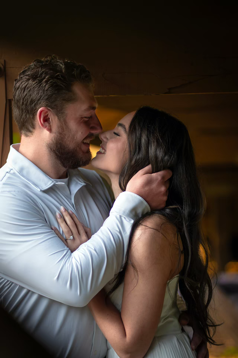 A couple shares a romantic embrace, smiling and about to kiss, with the woman wearing an engagement ring.