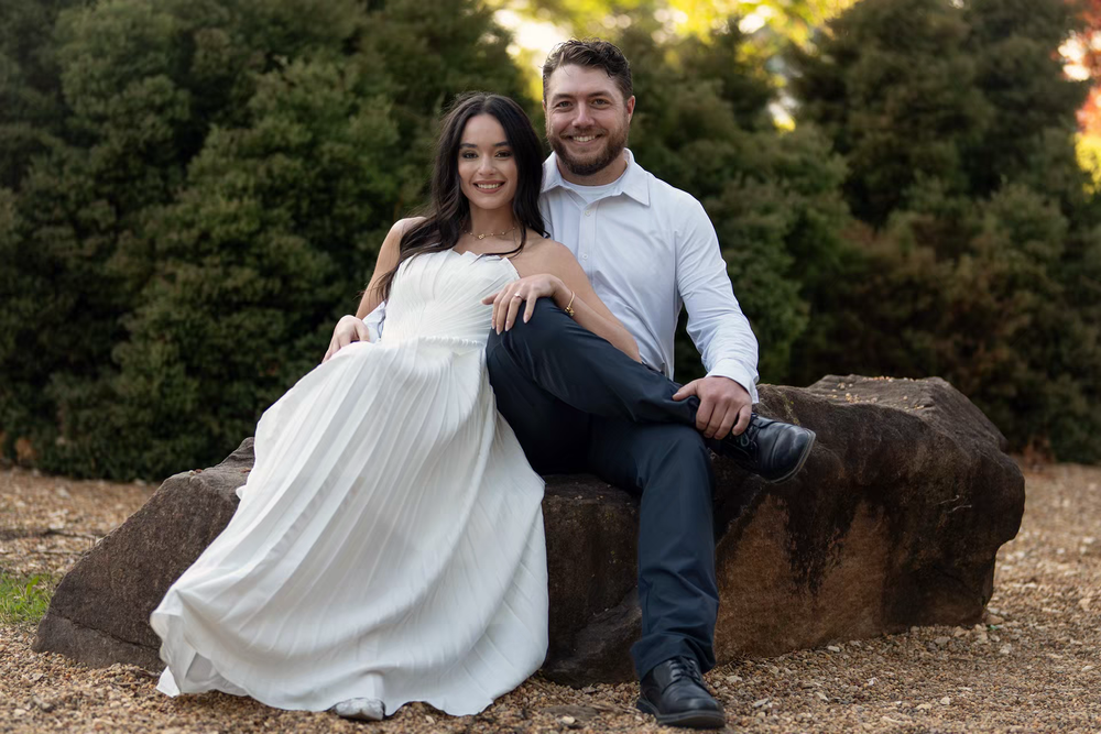 Happy couple posing on a rock in a garden, woman in white dress and man in white shirt smiling together.