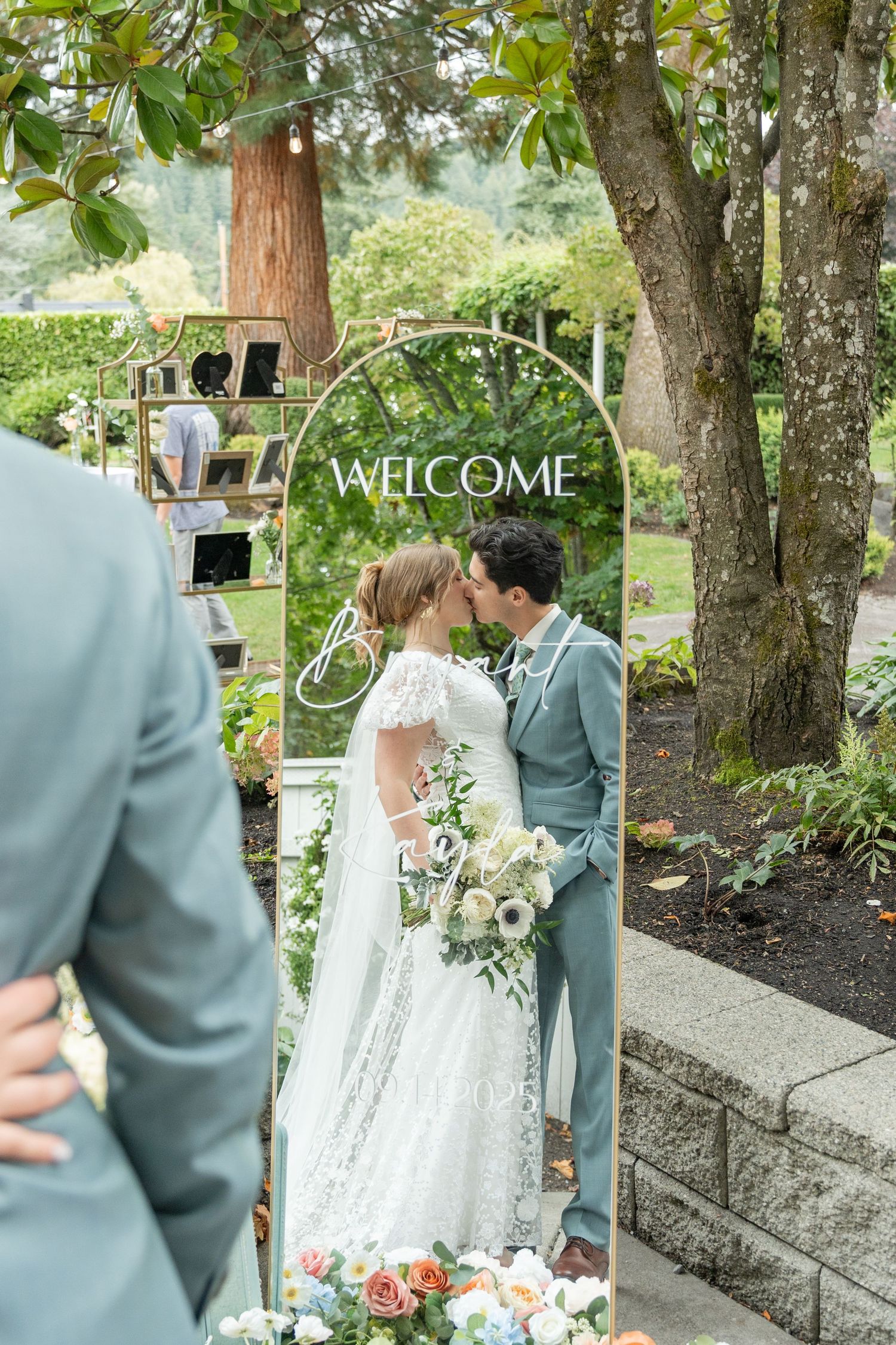 Bride and groom walking hand in hand on lush green lawn with tall evergreen trees and sunlit pond backdrop.