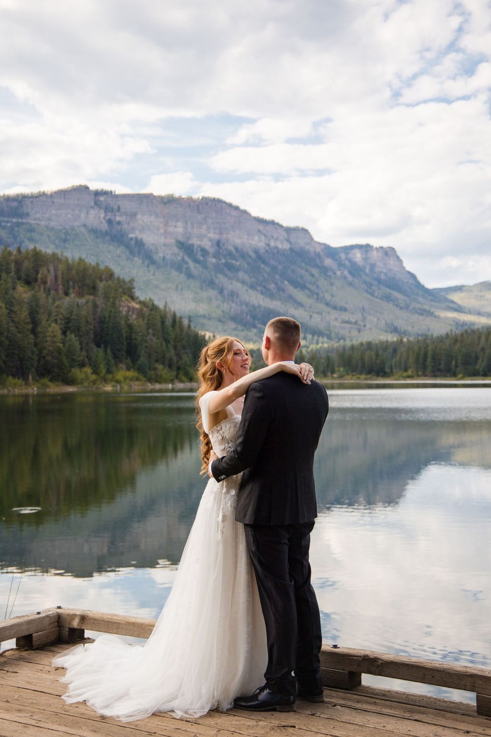 Bride and groom embrace on a wooden dock with a serene mountain lake and forested peaks reflected in calm water behind them.