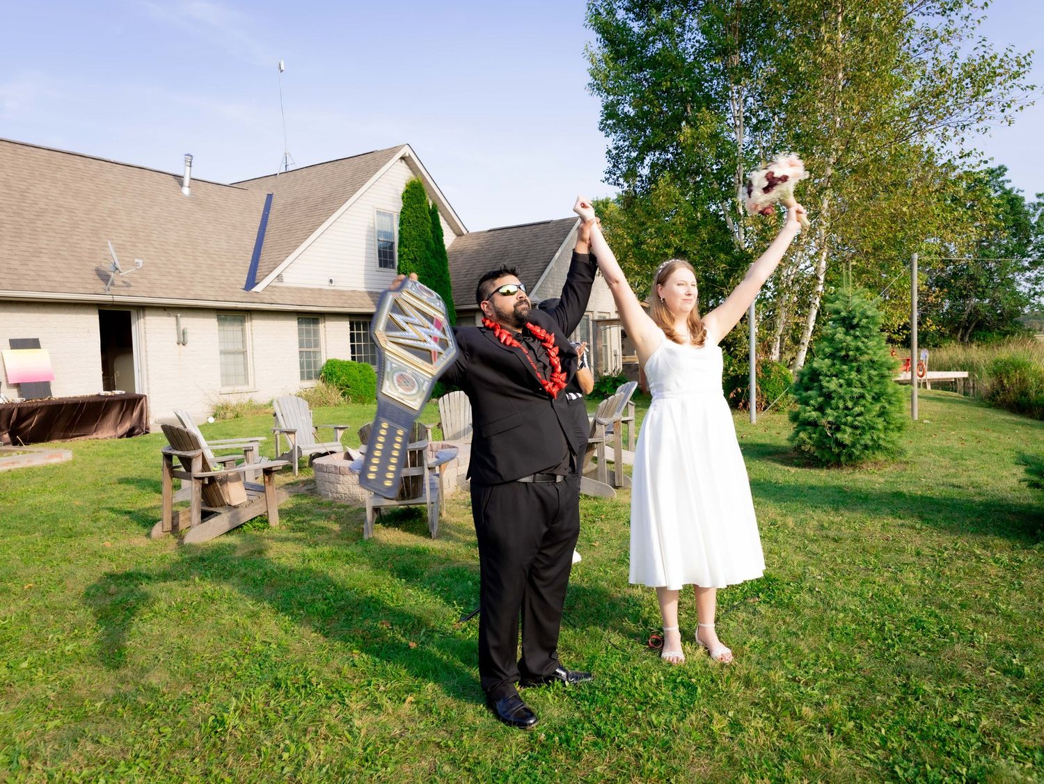 Joyful newlyweds celebrate outdoors in backyard, bride in white dress holding bouquet aloft, groom in black suit with red lei.