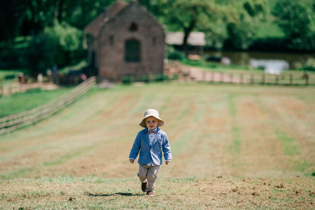 Young child in blue shirt and sun hat running across a green farm field with rustic barn in background.