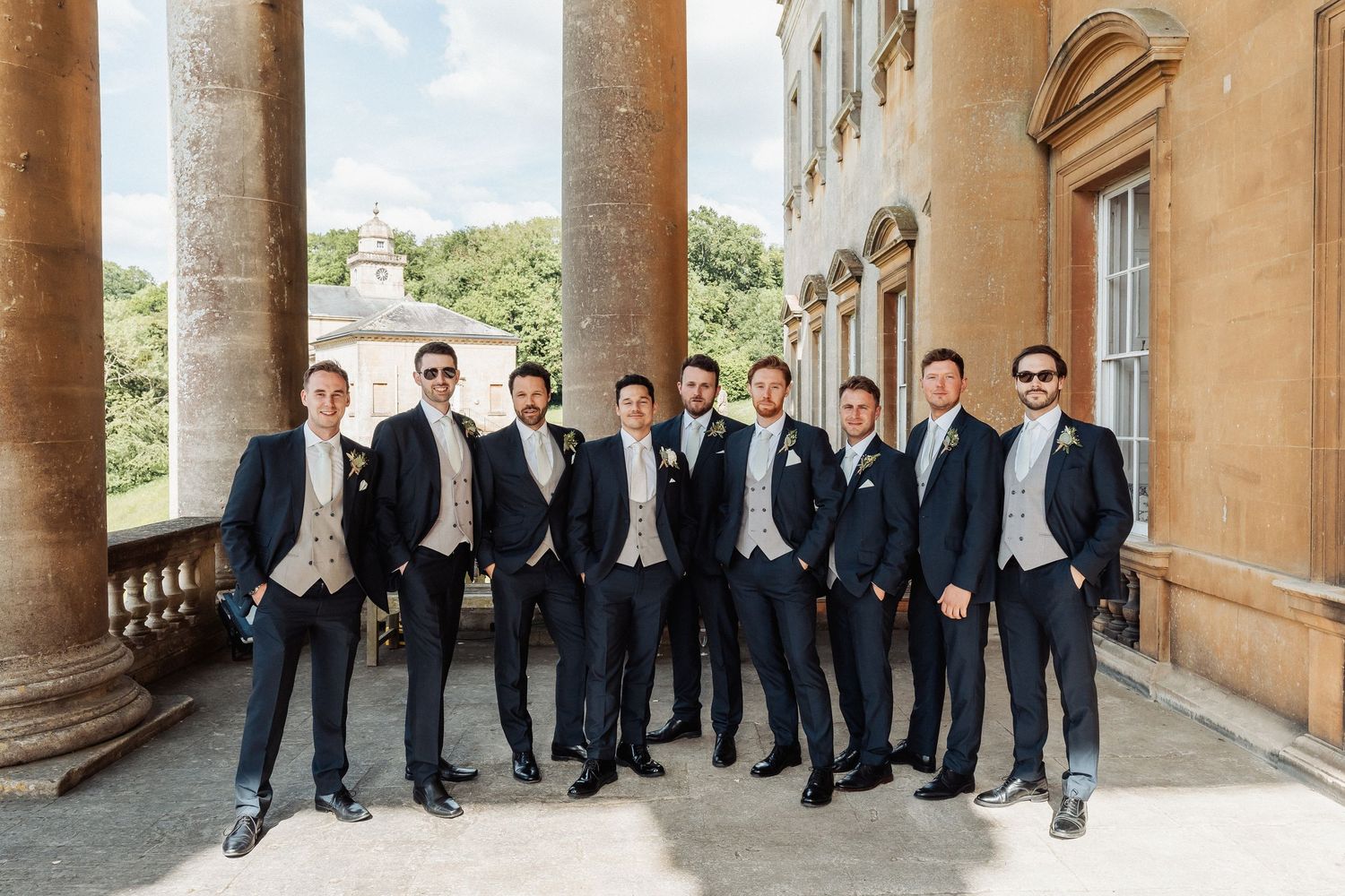 Groom and groomsmen in navy suits and cream vests pose at a grand stone venue with columns on a sunny wedding day.