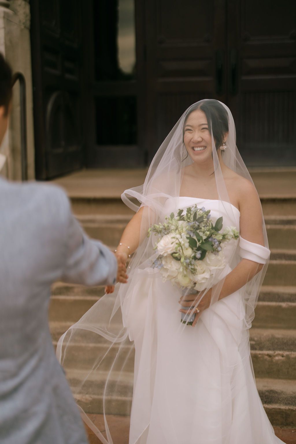 Smiling Asian bride in white strapless gown and veil holds bouquet on church steps, reaching toward groom.