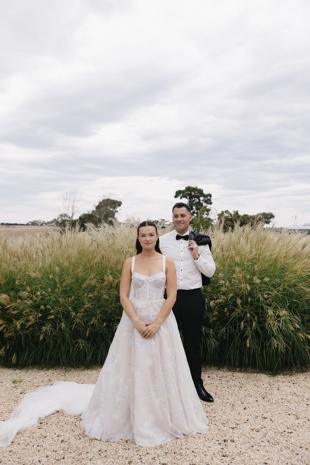 Bride in white lace gown and groom in black tuxedo pose before tall ornamental grasses on a cloudy day.