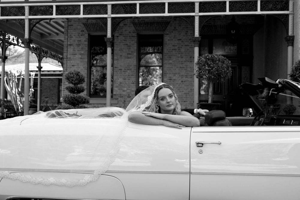 Bride in wedding veil leaning on classic white convertible car outside brick building, black and white photo.