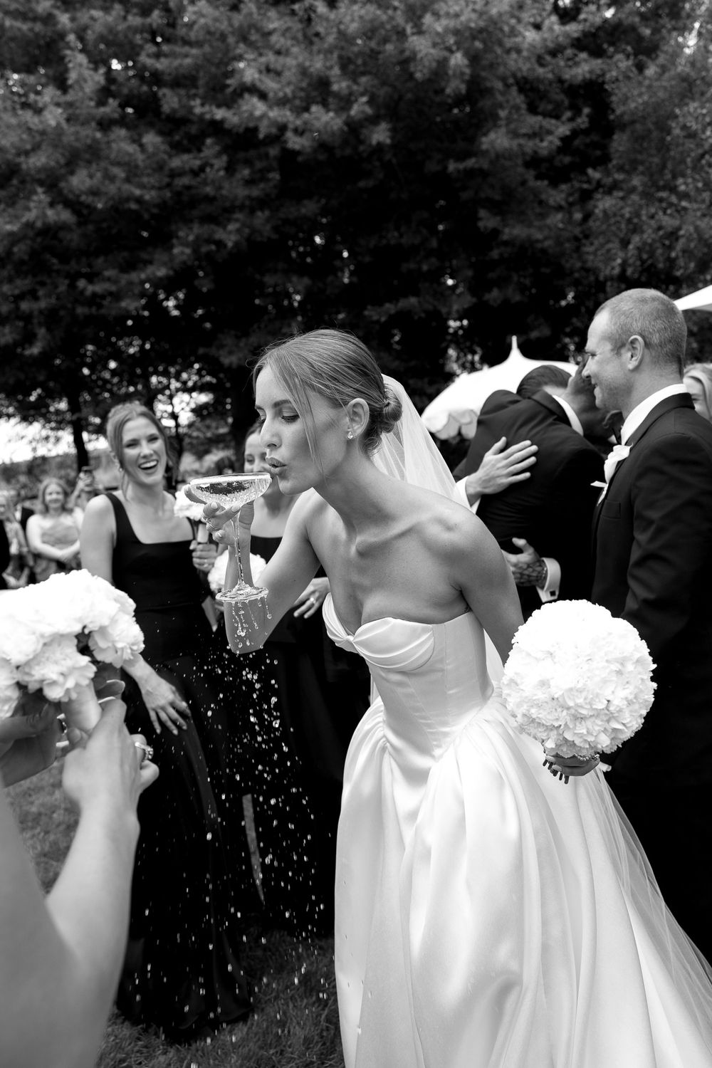 Bride in strapless gown sipping champagne while holding white bouquet, surrounded by guests at outdoor wedding reception.