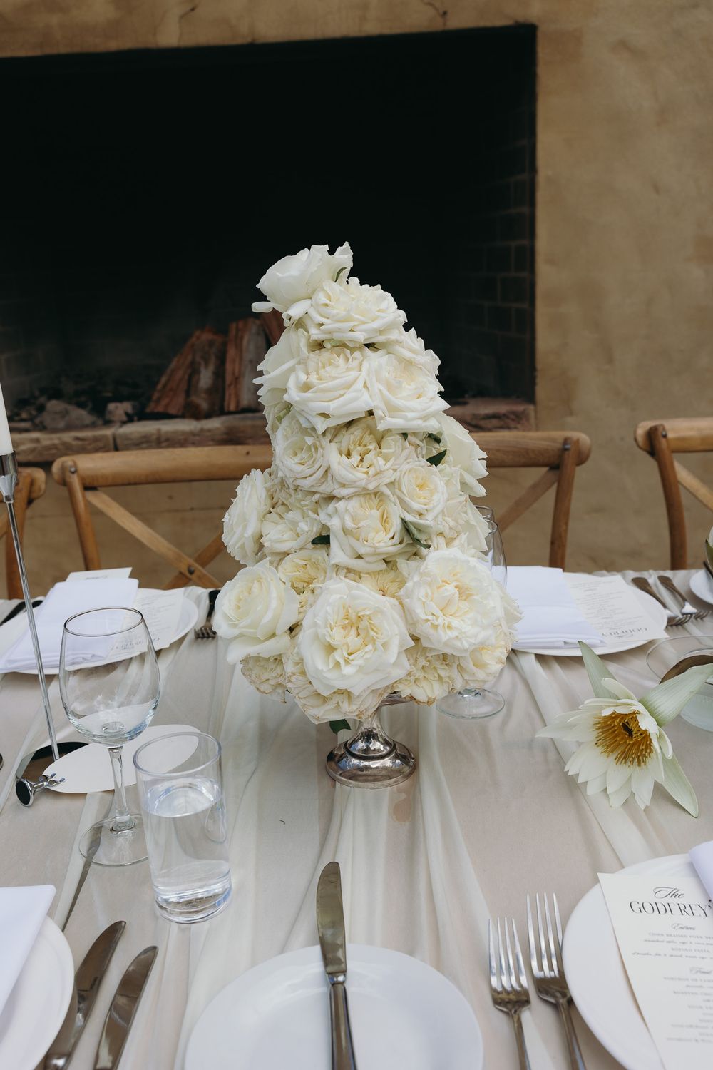 Elegant white rose centerpiece in silver vase on wedding reception table with fireplace backdrop.