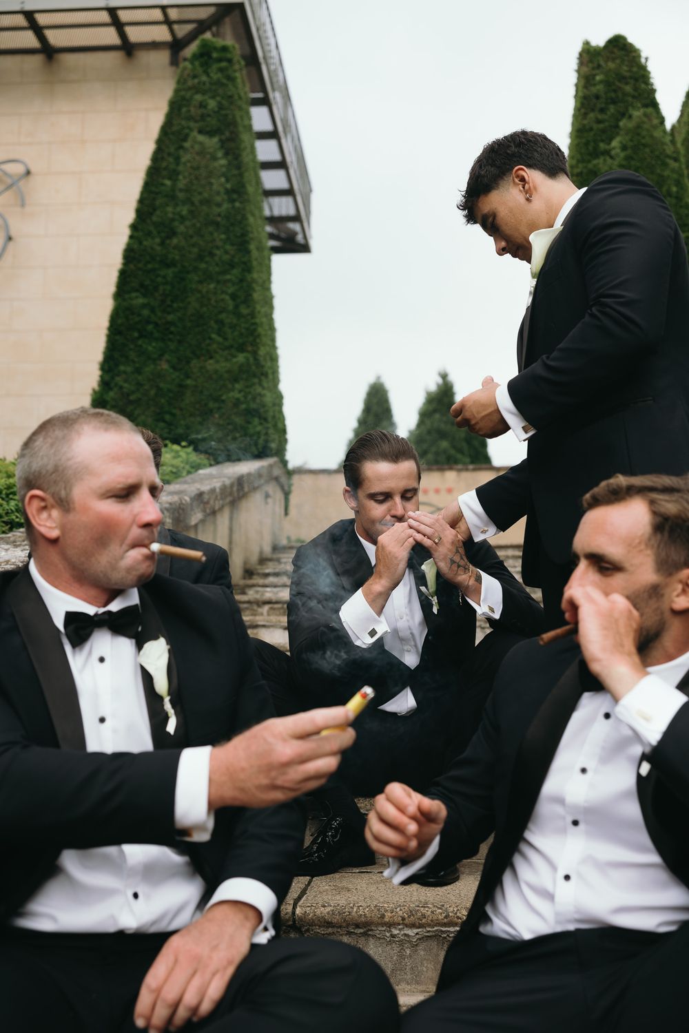 Groomsmen in black tuxedos smoking cigars outdoors on stone steps at a wedding celebration.