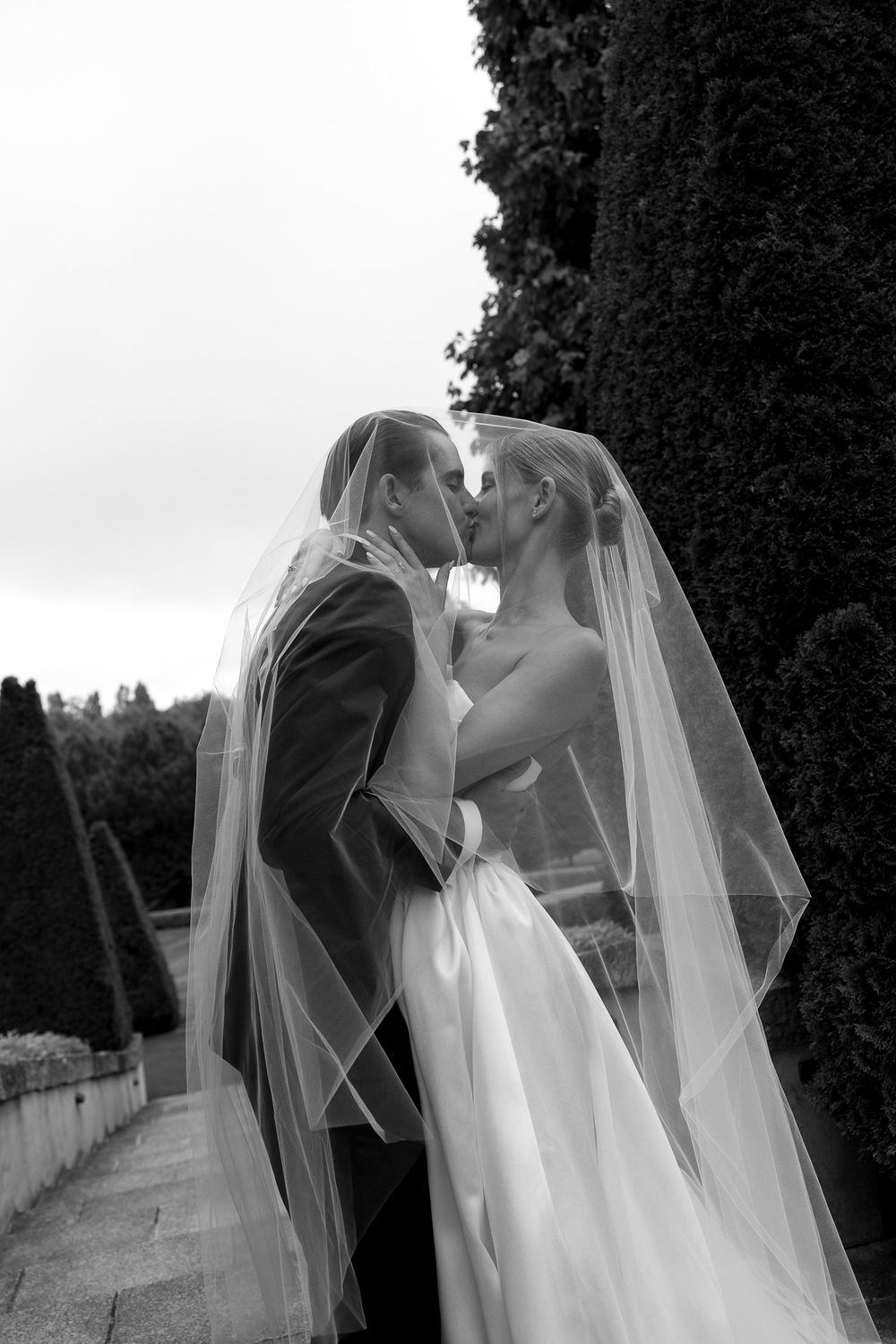 Black and white photo of bride and groom kissing under a wedding veil outdoors.