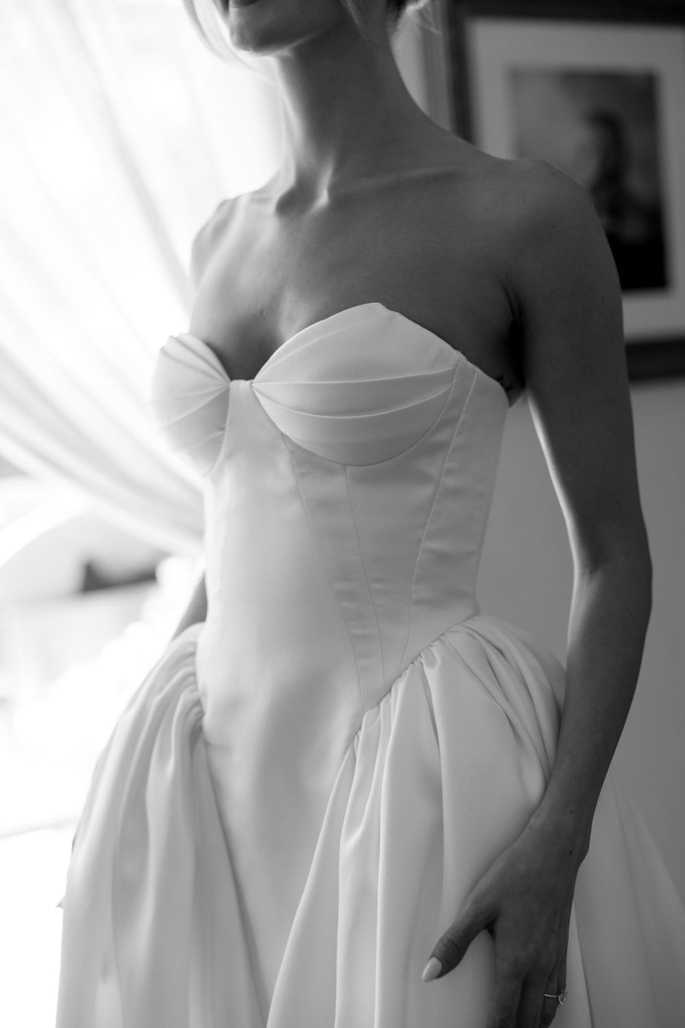 Black and white photo of a bride in a strapless sweetheart wedding gown standing by a window.