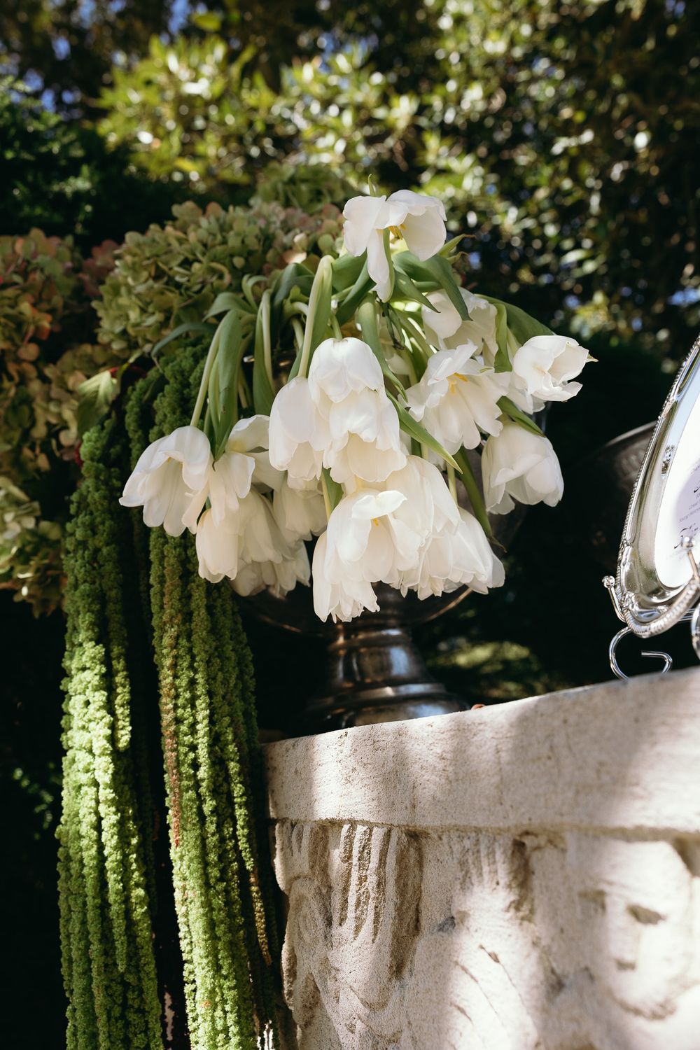 White tulips drooping in a bronze vase on a stone pedestal, surrounded by lush greenery outdoors.