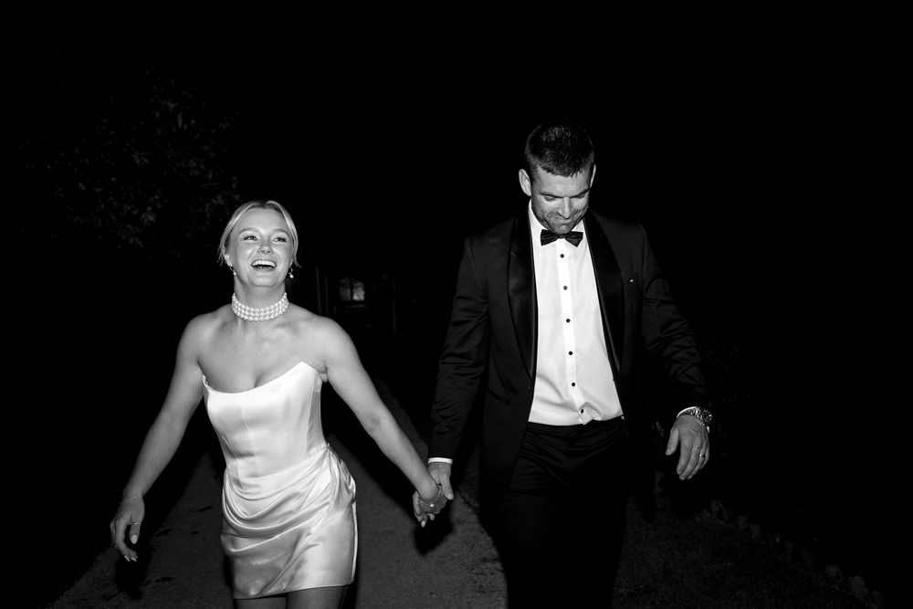 Black and white photo of a laughing bride in a short dress and groom in a tuxedo holding hands at night.