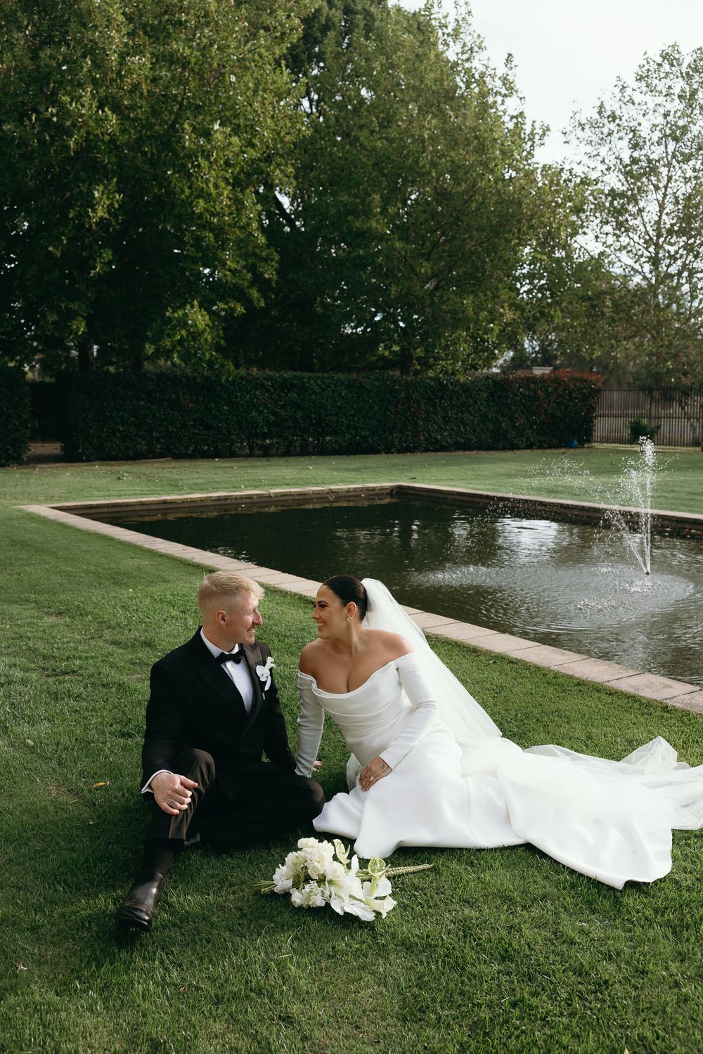 Bride and groom sitting on green grass by a fountain in a garden, wearing white gown and black tuxedo.