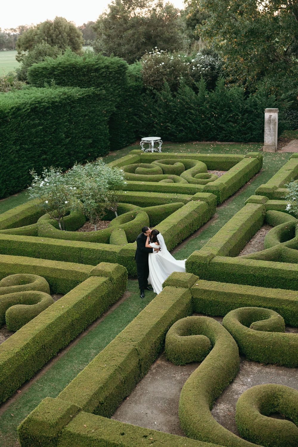 Bride and groom embracing in an ornate formal garden maze with sculpted hedges at their wedding venue.