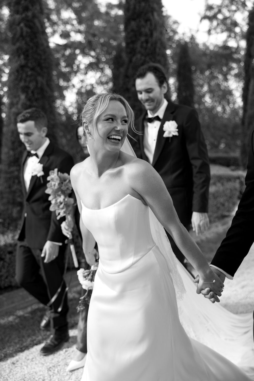 Black and white photo of a laughing bride in a strapless gown holding hands with groom in tuxedo at outdoor wedding.