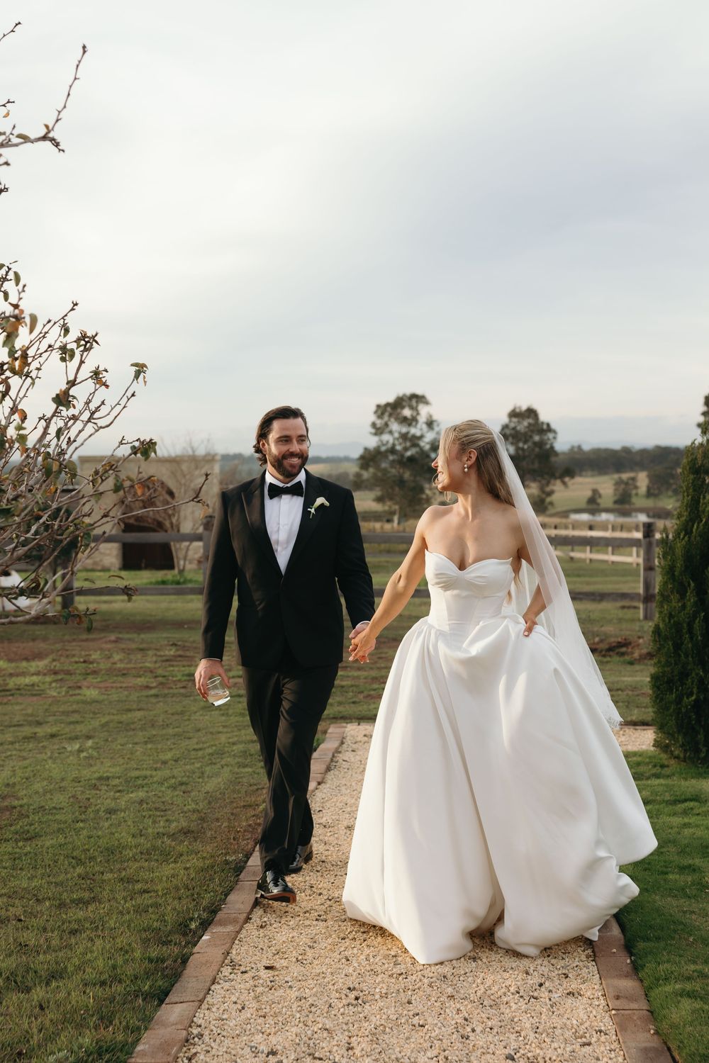 A bride and groom walk hand-in-hand along a garden path at an outdoor wedding venue, smiling at each other.