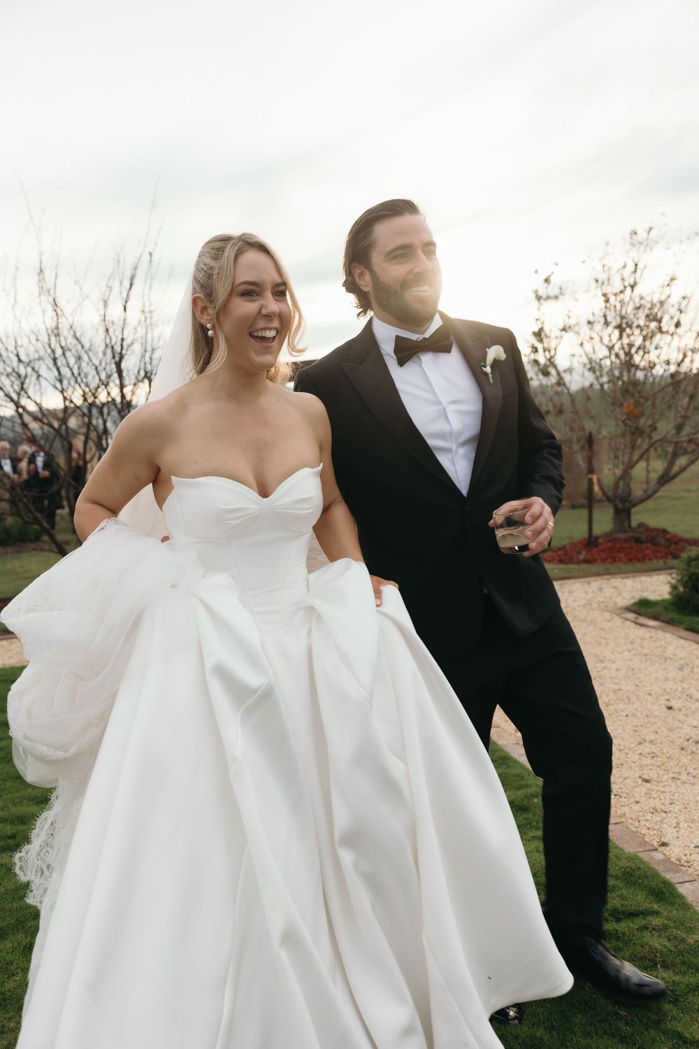 A joyful bride in a white strapless gown and groom in black tuxedo walk together outdoors after their wedding ceremony.