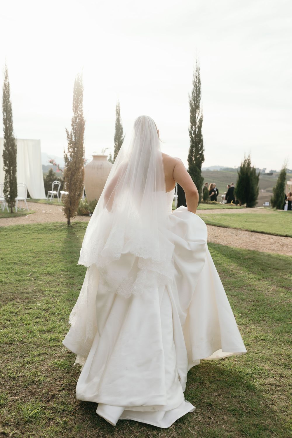 Bride in a flowing white ball gown and veil walks through a garden toward tall cypress trees at an outdoor wedding.