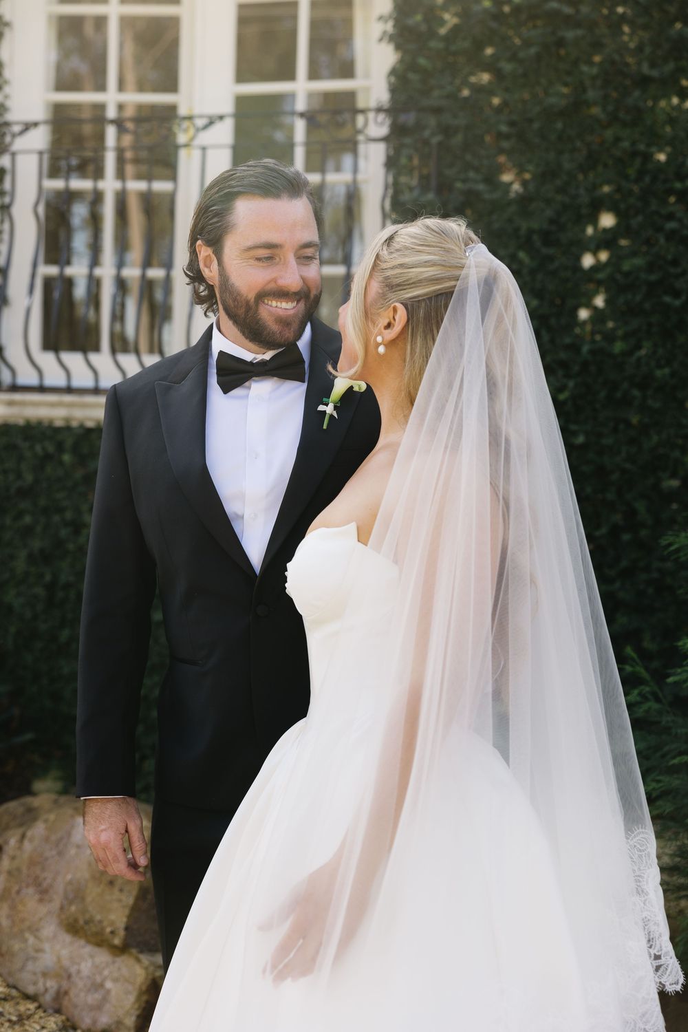 A smiling groom in a black tuxedo gazes at his bride in a white strapless gown and veil outdoors.