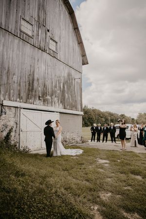 Bride and groom share a moment beside a rustic barn while guests and photographer capture the scene.
