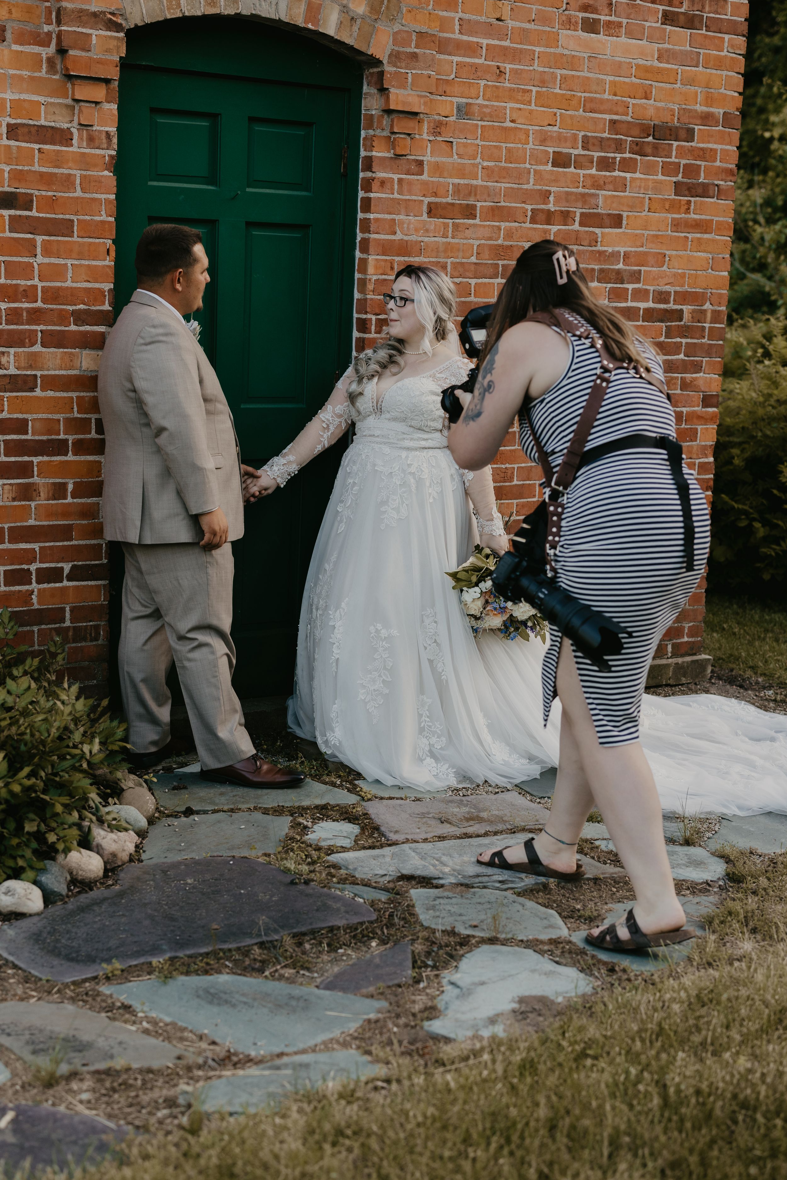 Wedding photographer directs bride and groom posing by a green door on a brick building exterior.
