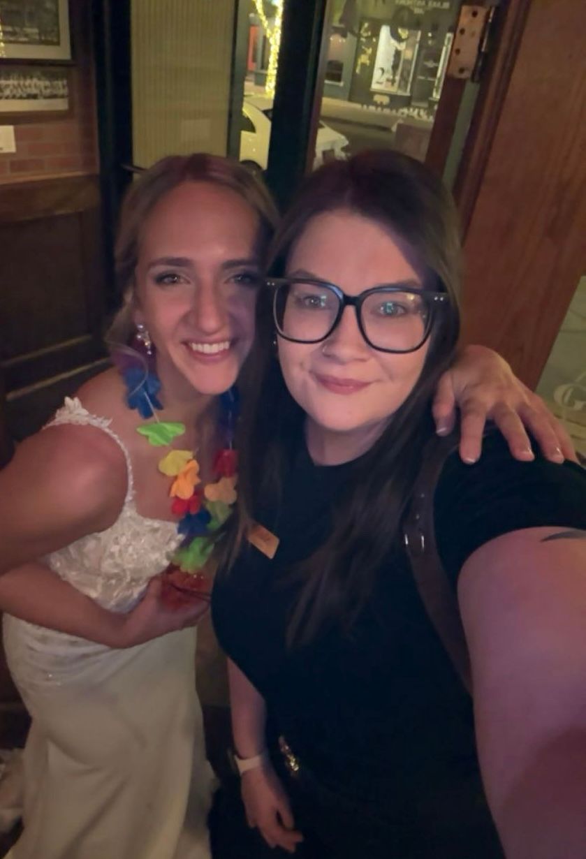 Wedding photographer takes a selfie with a smiling bride wearing a floral lei at a reception.