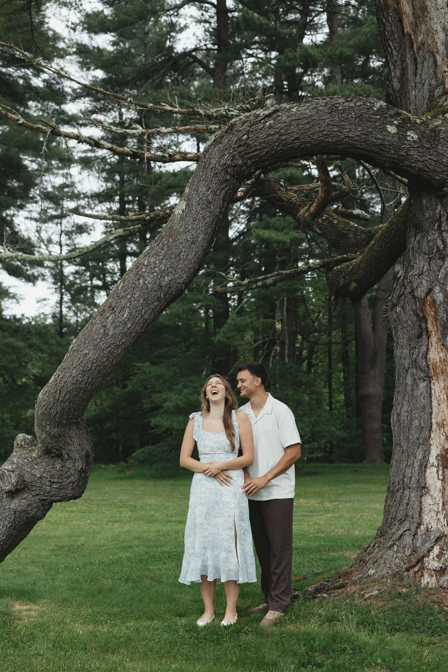 Happy couple laughing together under a large arching tree in a lush green park, woman in floral dress touching her baby bump.