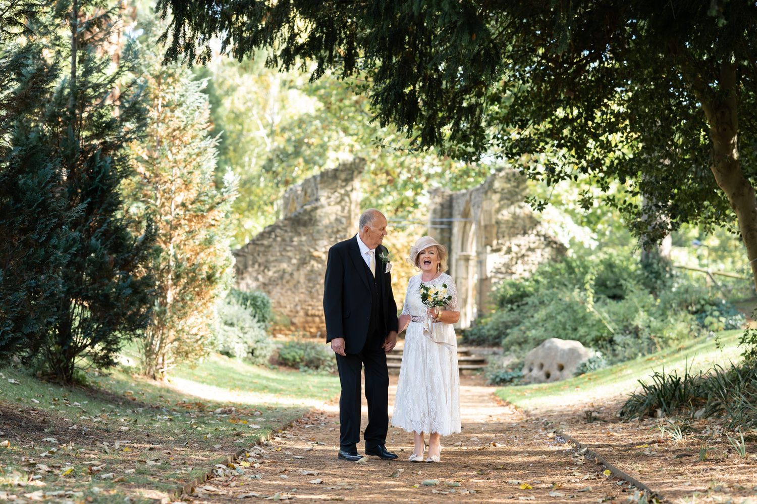 Mature bride and groom walking hand in hand along a sunlit tree-lined garden path in Abingdon.
